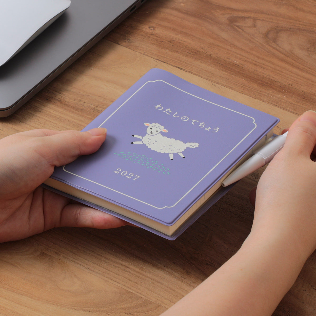 Hands holding a Purple 2027 sheep-cover planner on a wooden desk, with a white pen and laptop nearby.