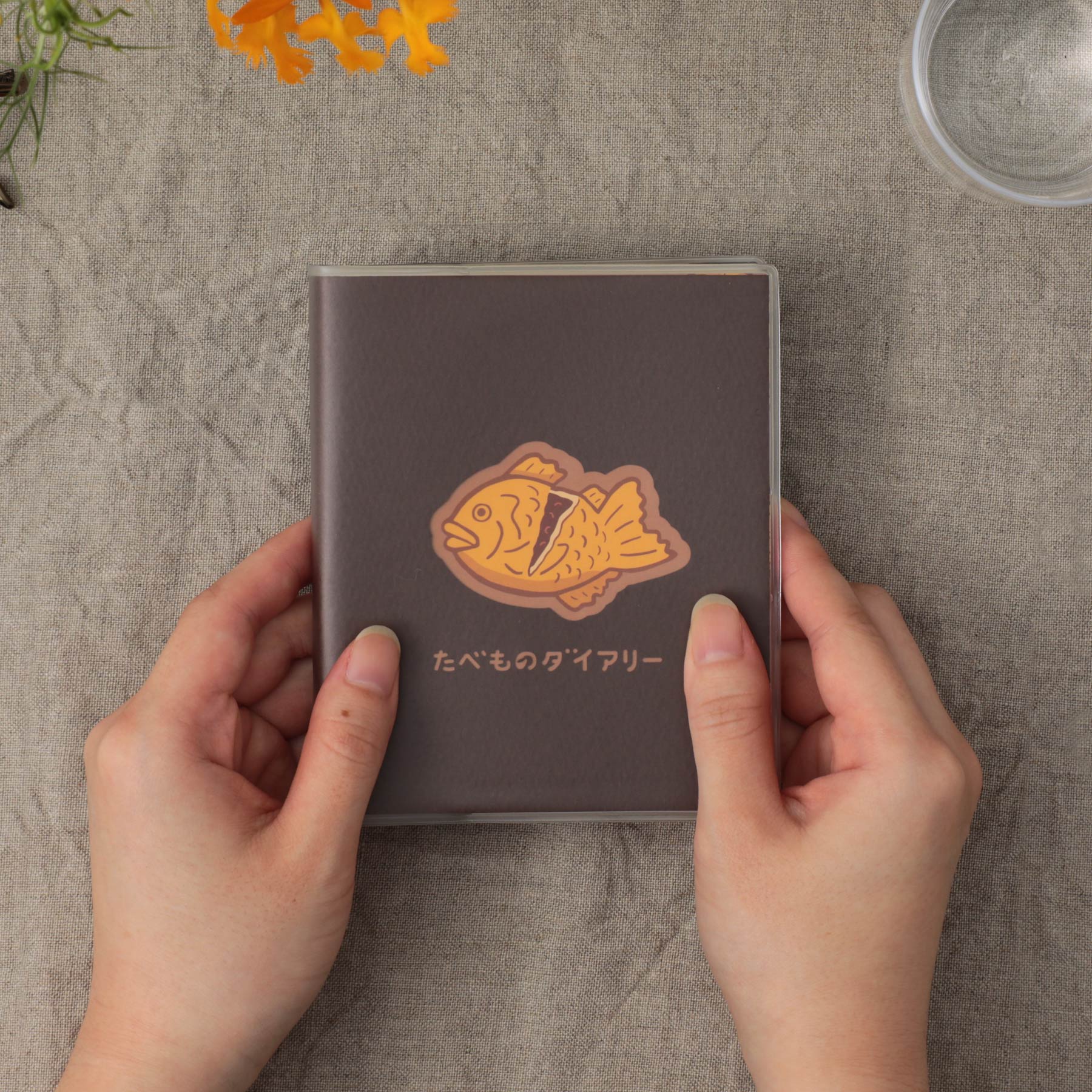 Two hands holding a Brown food diary planner with a taiyaki fish illustration on the cover, over linen fabric.