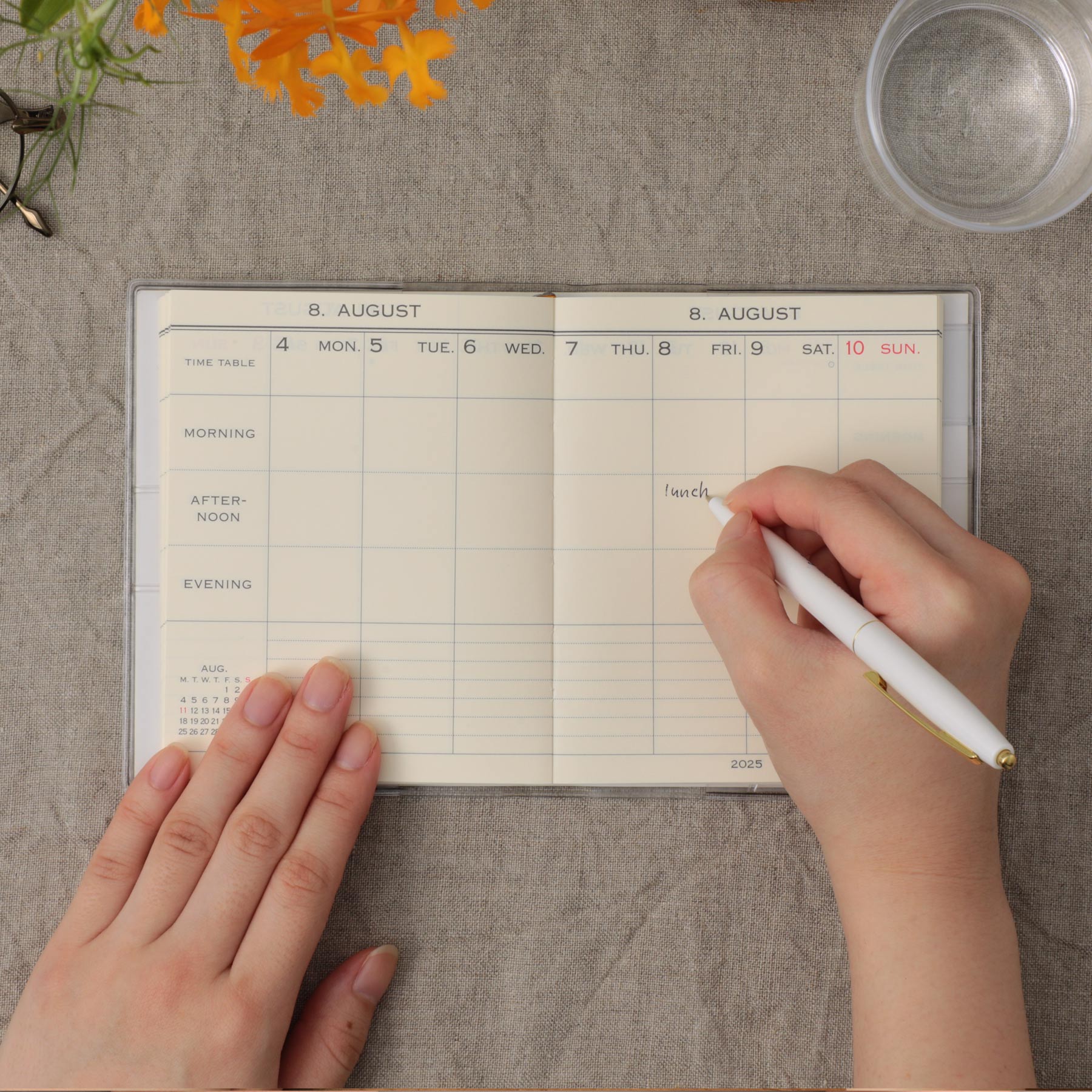 Hands writing "lunch" with a white pen in an open weekly planner with time-table grid on linen fabric.