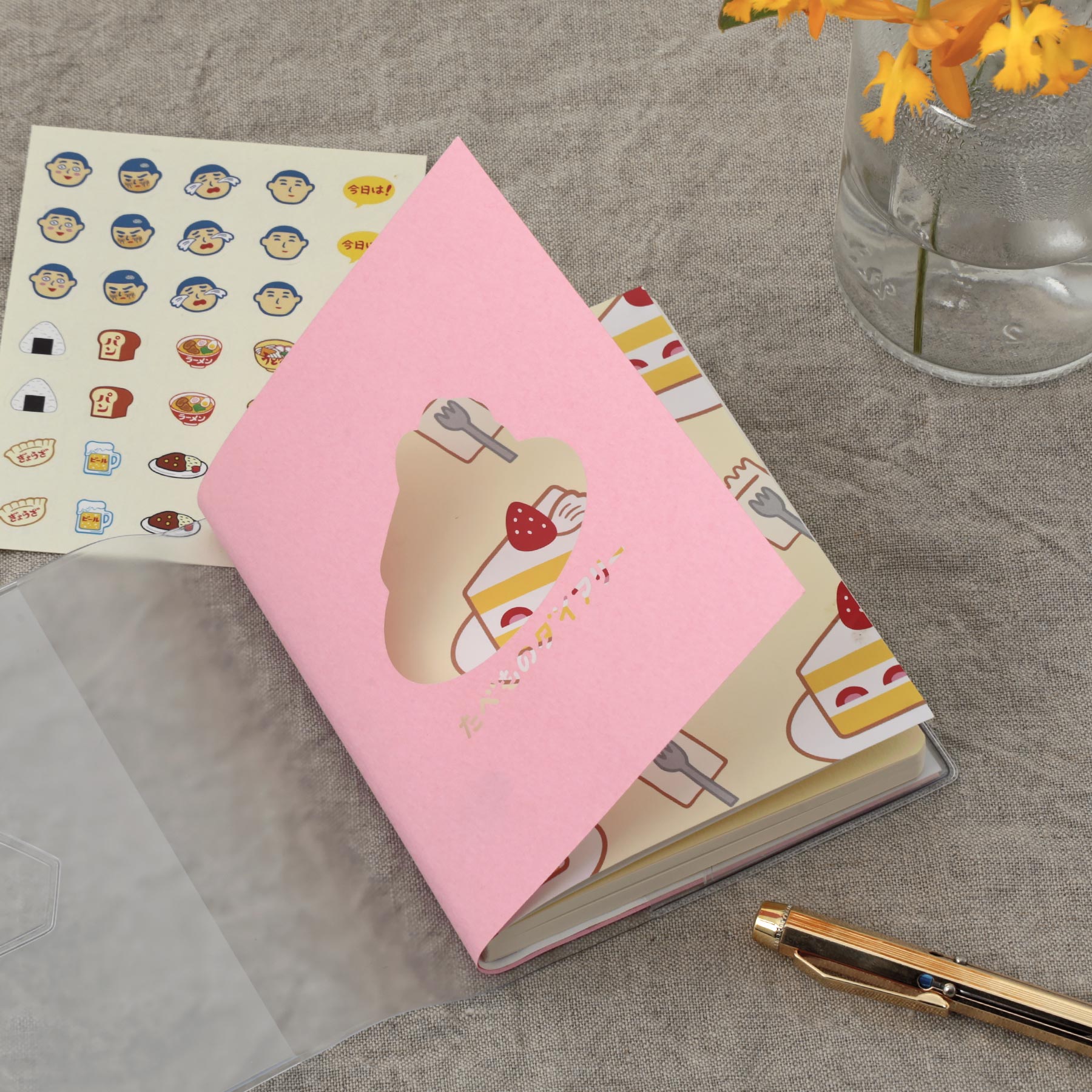 Planner with Pink die-cut cover sliding over a strawberry cake pattern, beside food stickers and a gold pen.