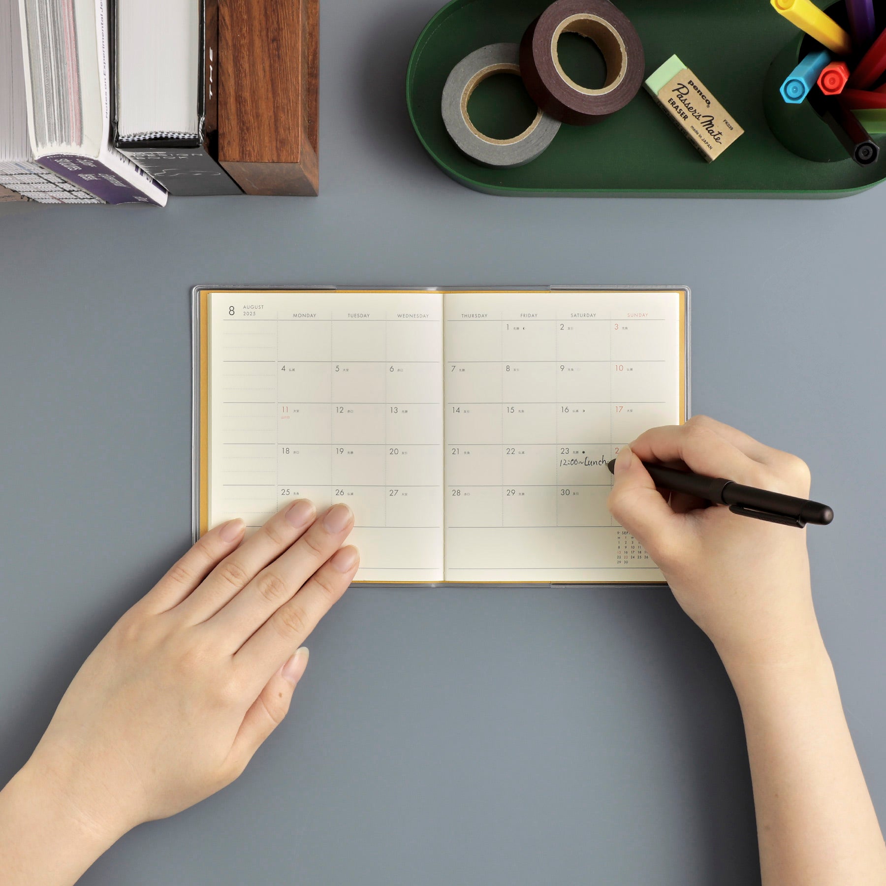 Hands writing a lunch appointment in an open Nähe monthly planner spread beside pens and tape
