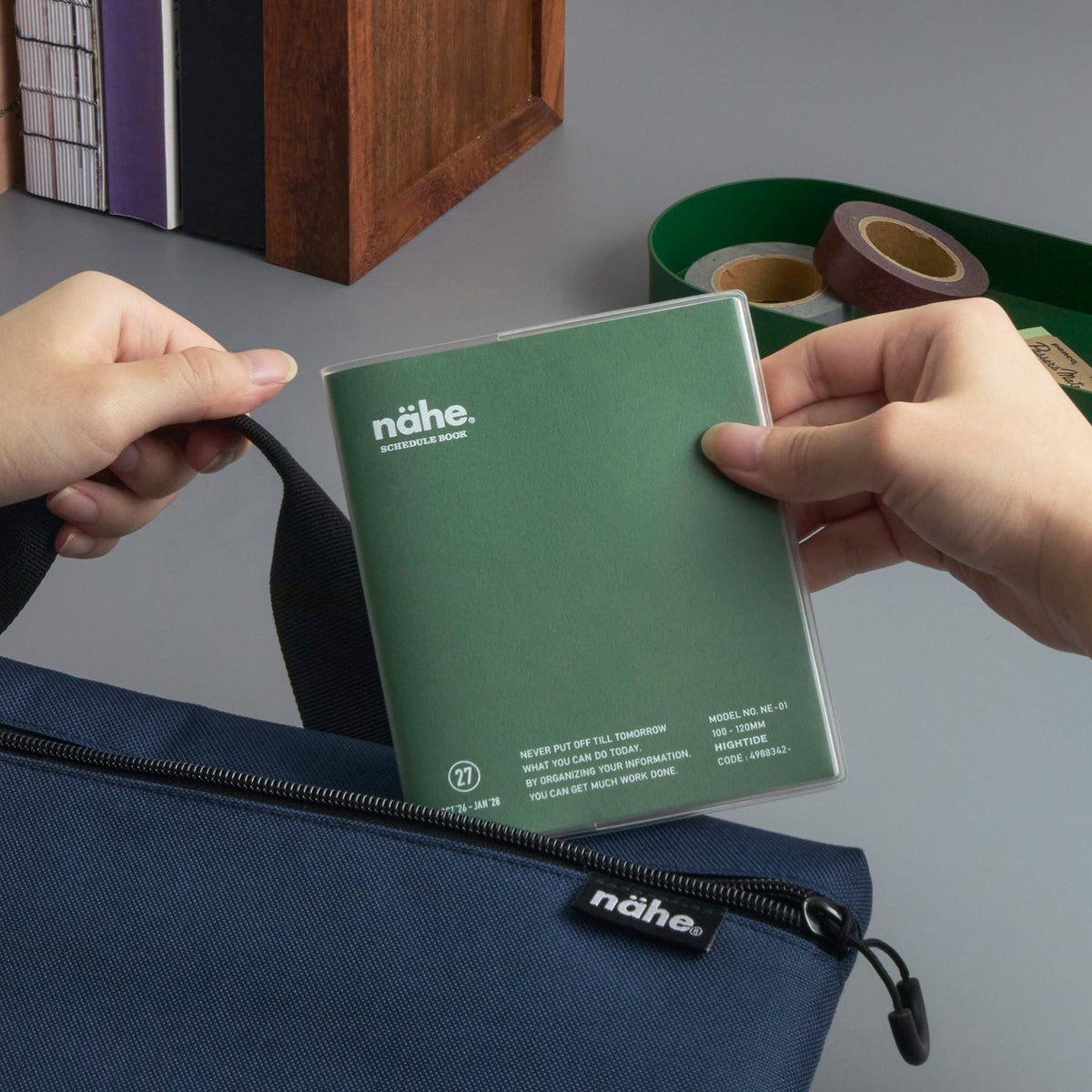 Hands lifting a Green Nähe square planner in a clear sleeve out of a navy zip pouch beside washi tape.