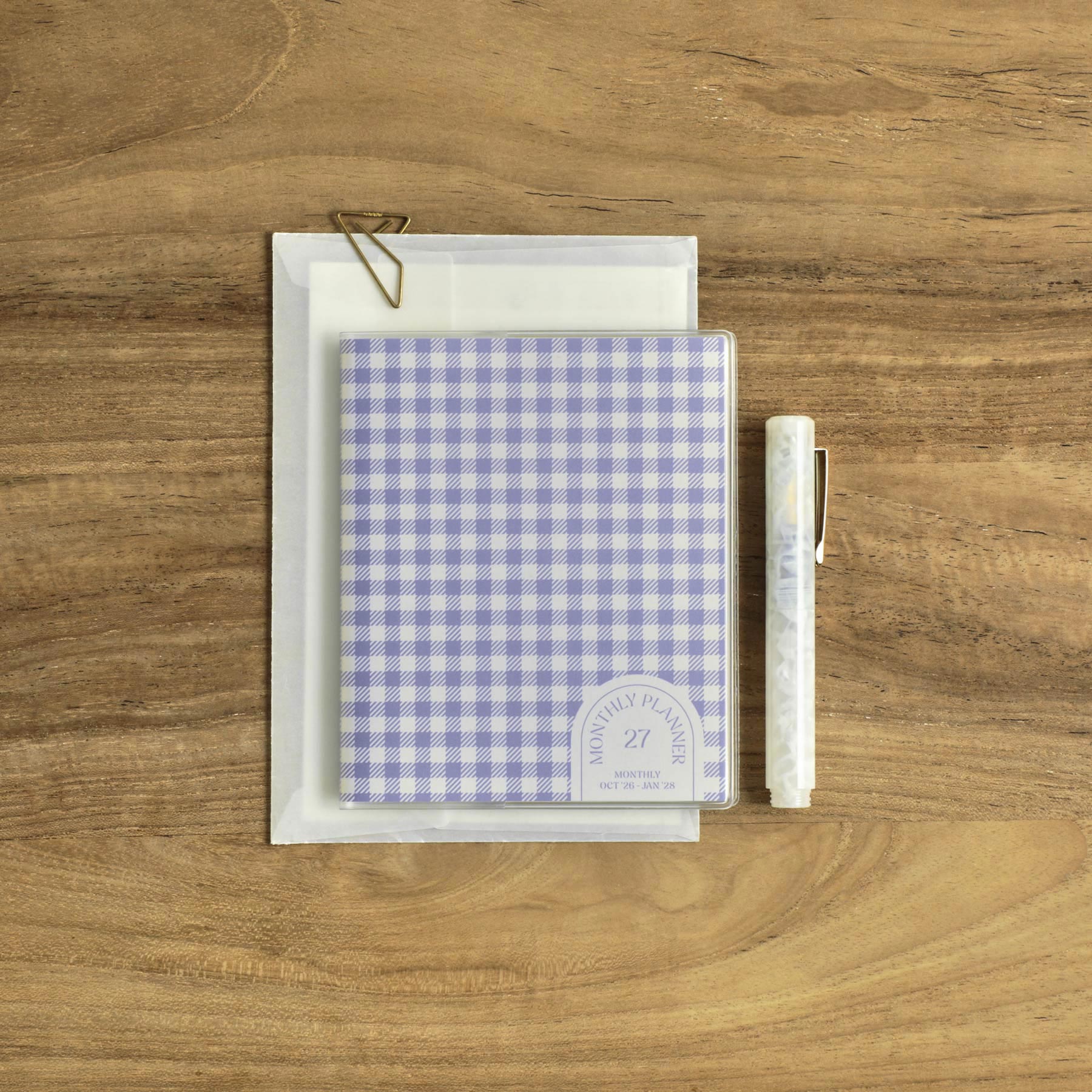 Lavender gingham check monthly planner on an envelope with a gold clip and pen on a wood surface.