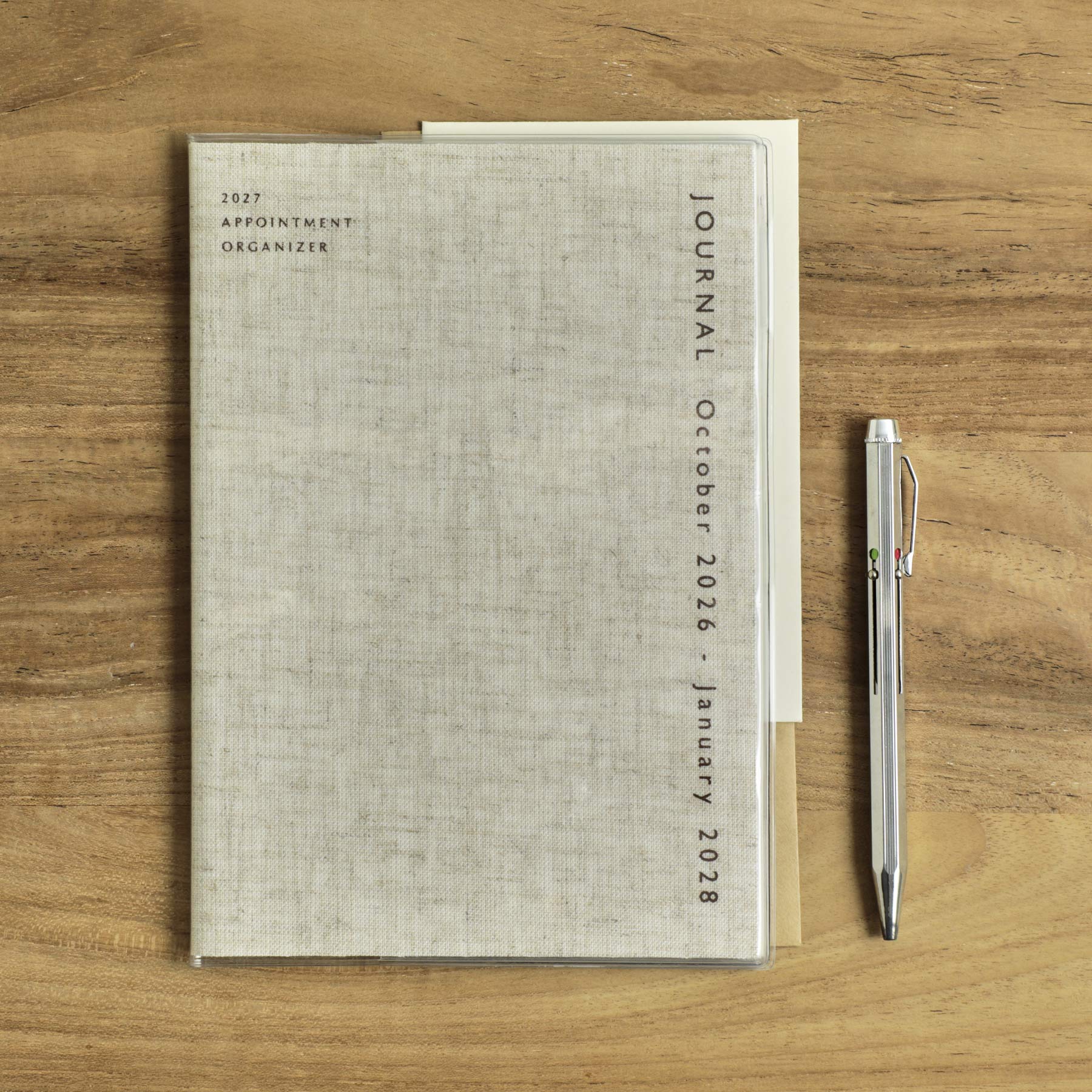 Hightide 2027 Coton A5 monthly planner in Linen with clear cover, beside a silver pen on wood.