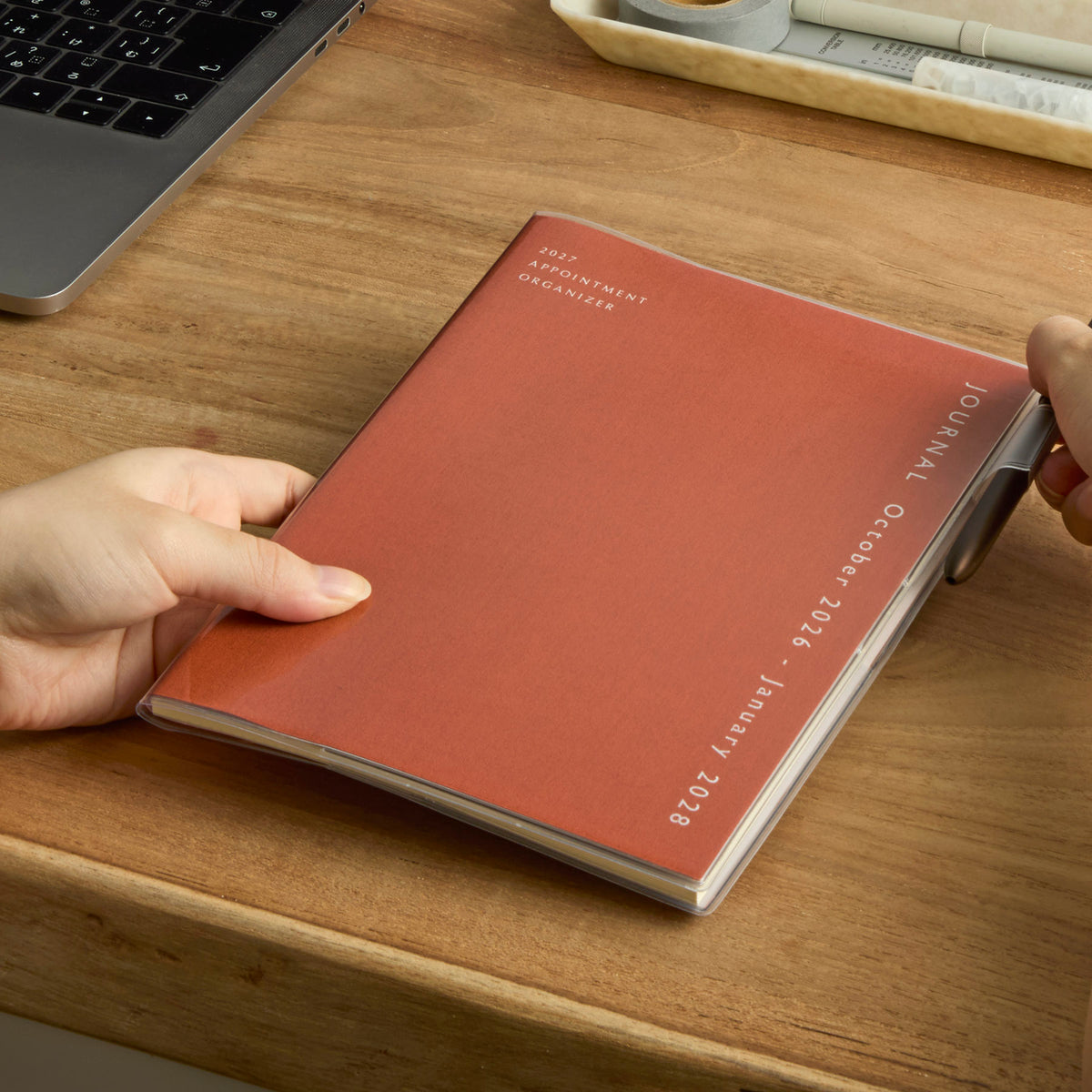 Hands holding an Orange fabric-covered Hightide planner with clear cover on a wooden desk beside a laptop.