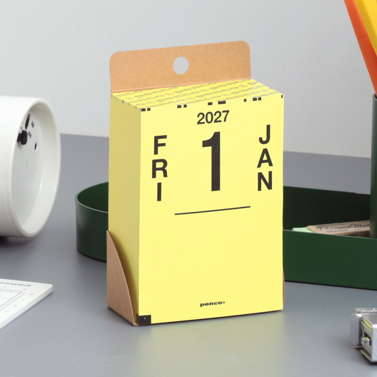 Penco 2027 daily calendar in yellow with kraft backing, showing FRI 1 JAN on a desk beside a green tray.