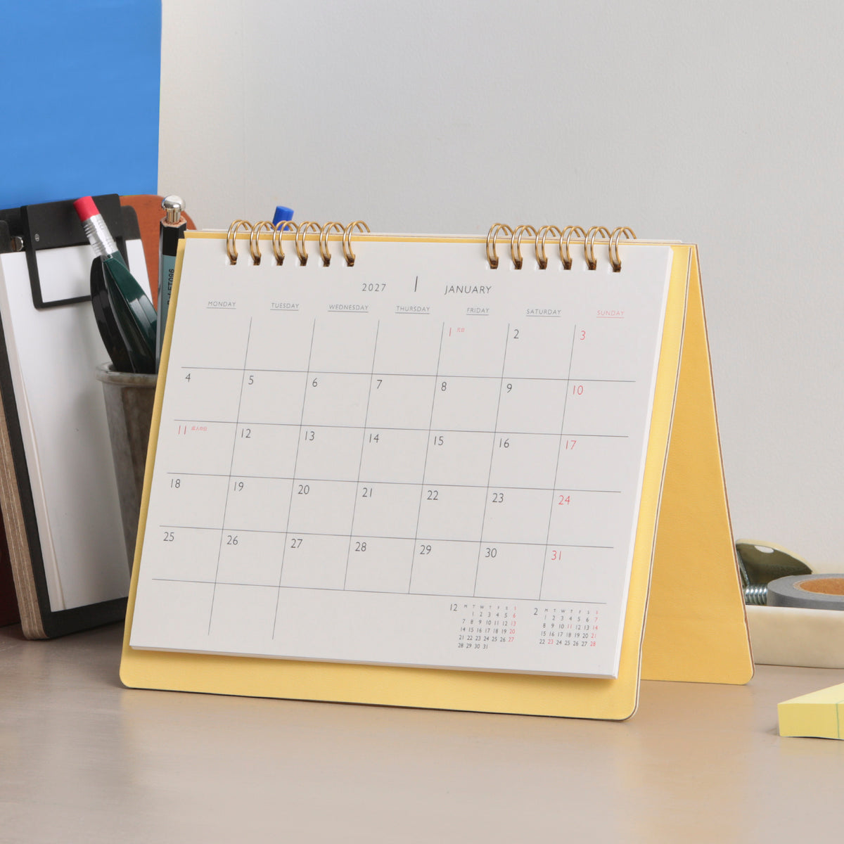 Light Yellow standing desk calendar with twin gold wire binding, open to a January 2027 monthly grid.