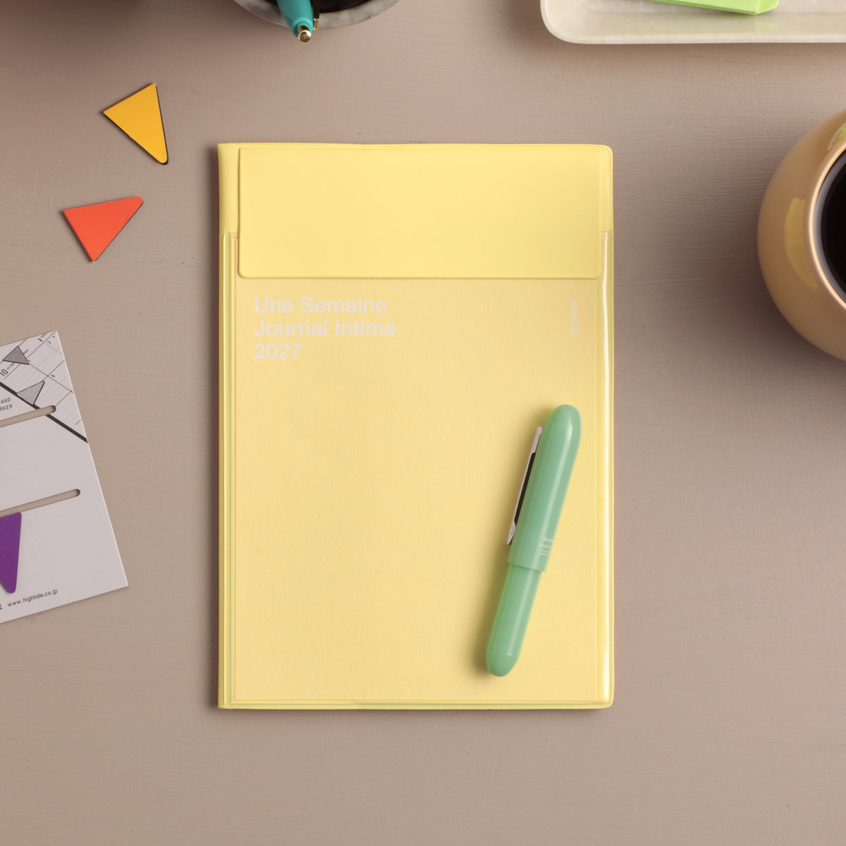 Yellow 2027 monthly planner with envelope-style pocket cover, styled on a desk with a mint pen and triangle clips.