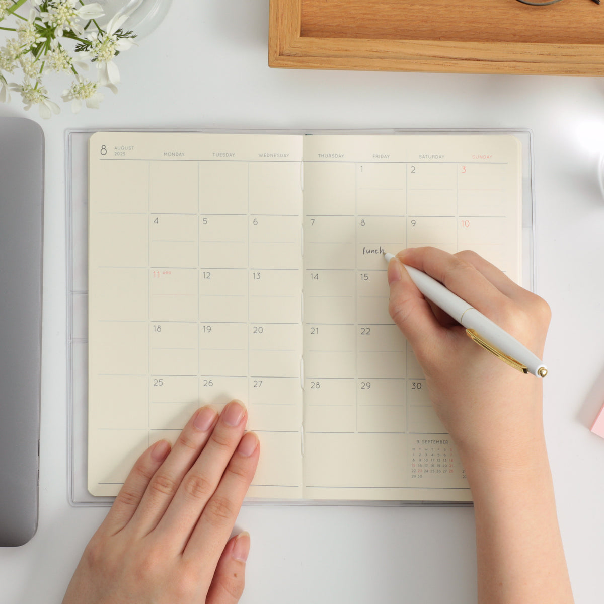 Hands writing "lunch" with a pen in an open monthly planner showing an August calendar spread.