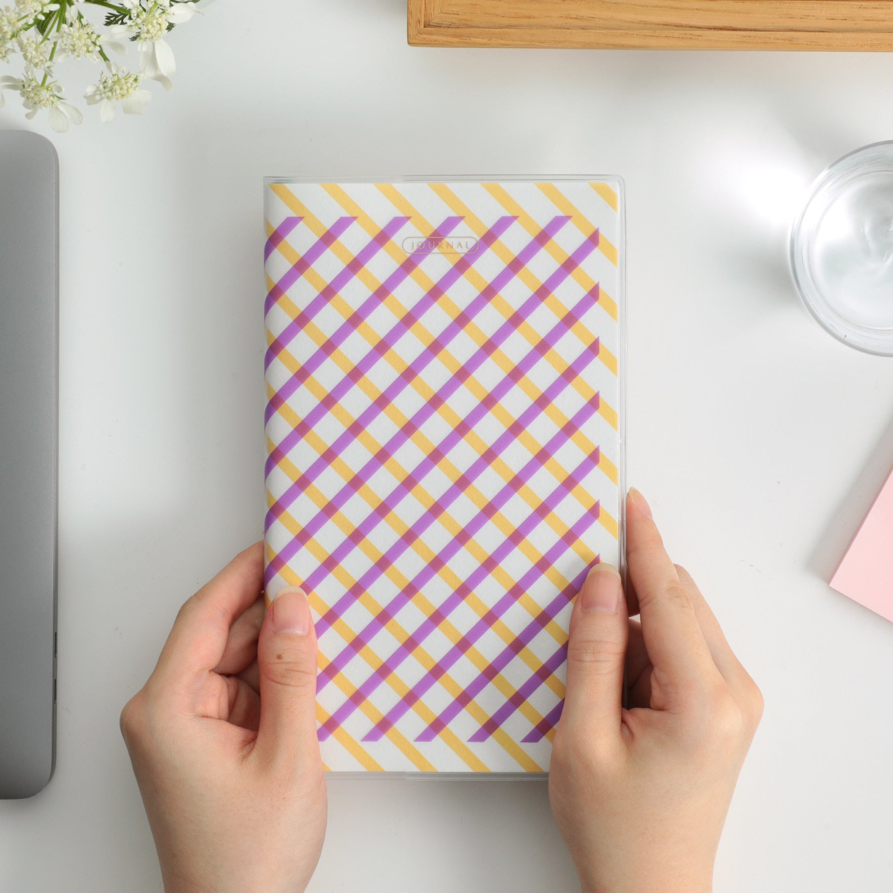 Hands holding a Hightide B6 slim planner with a Purple and yellow diagonal plaid cover on a white desk.