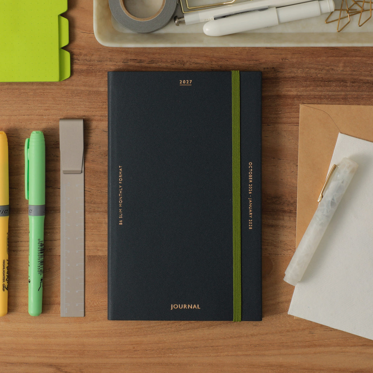 Navy 2027 Katy B6 Slim planner with green elastic band on a wooden desk with pens and a highlighter.
