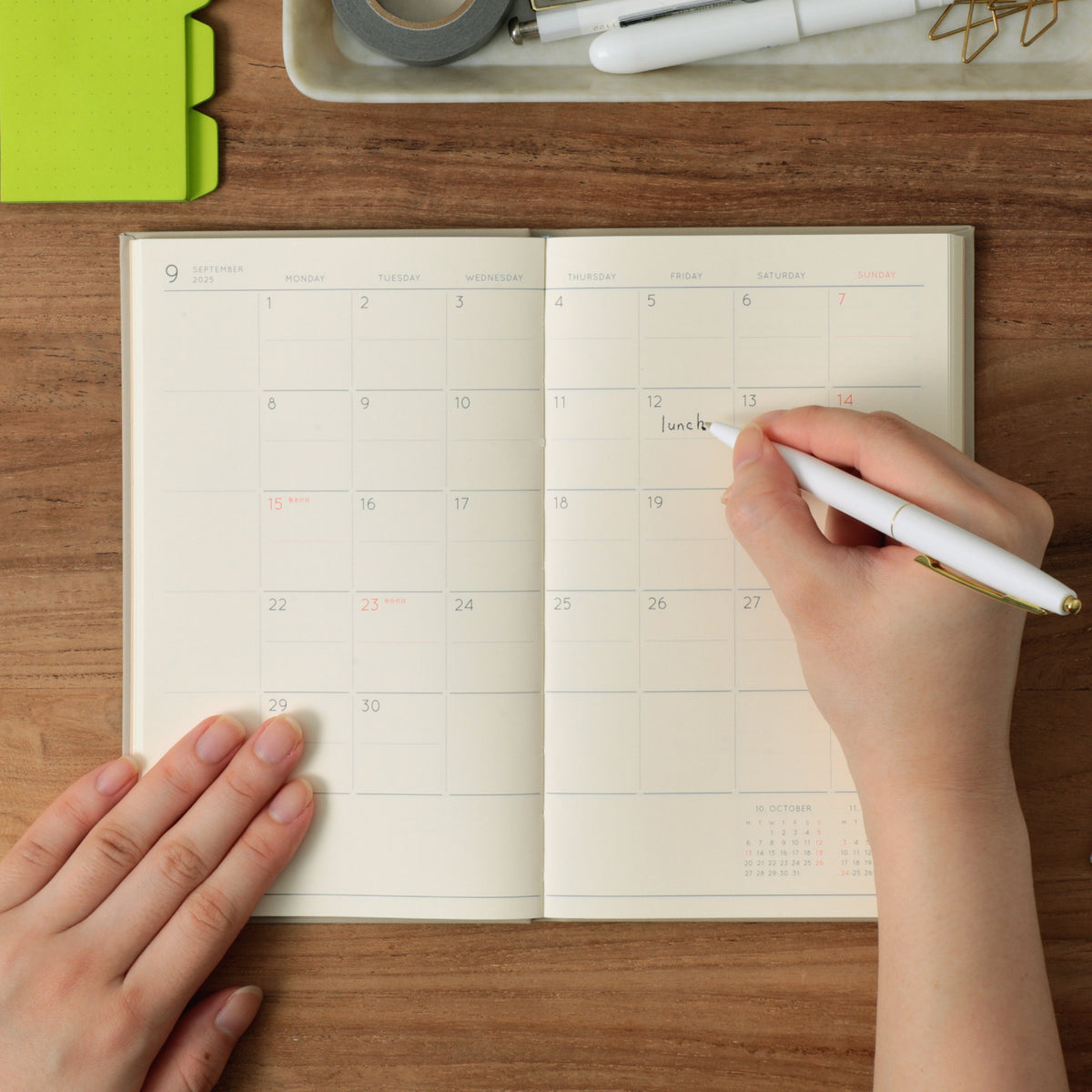 Hand writing "lunch" with a white pen on the open September monthly grid of a planner on a wooden desk.