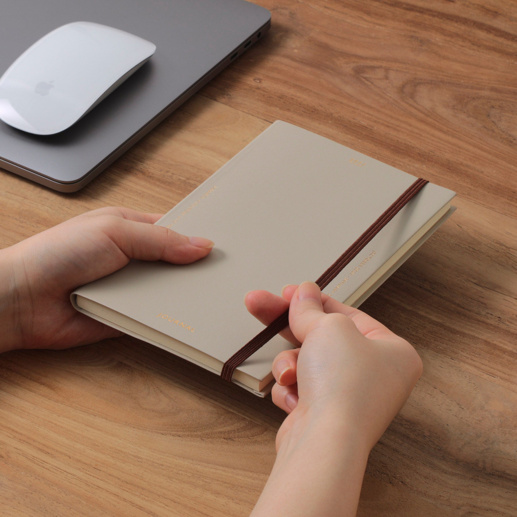 Hands holding a Beige Katy planner with a brown elastic band on a wooden desk beside a laptop and mouse.