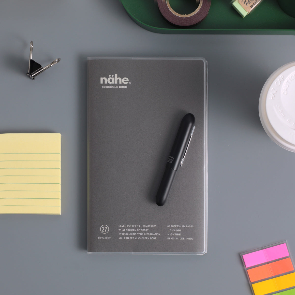 Gray Nähe schedule book in a clear cover with a black pen on top, beside sticky notes and clips.