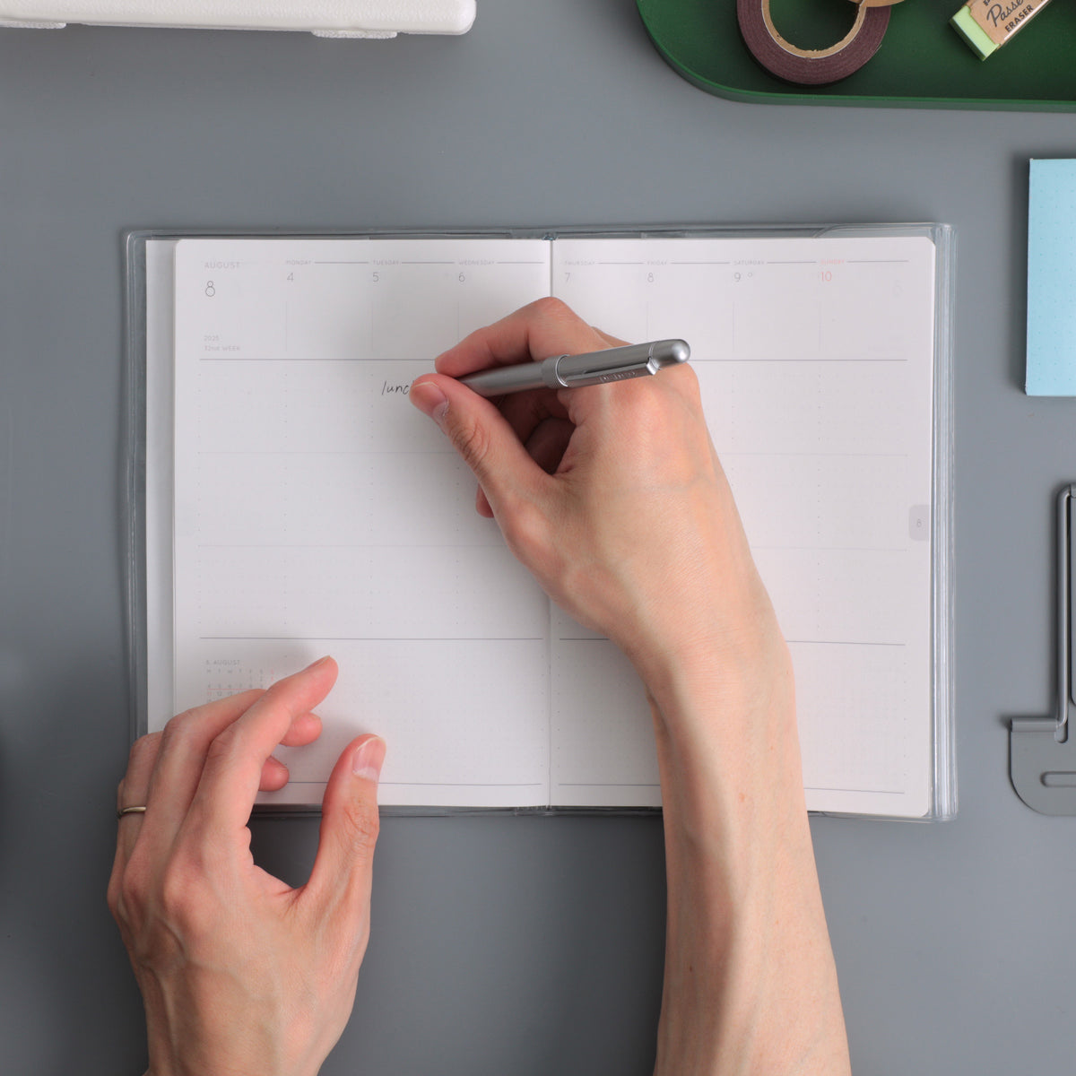 Hands writing with a silver pen in an open Nähe B6 weekly planner with a Clear cover on a grey desk.