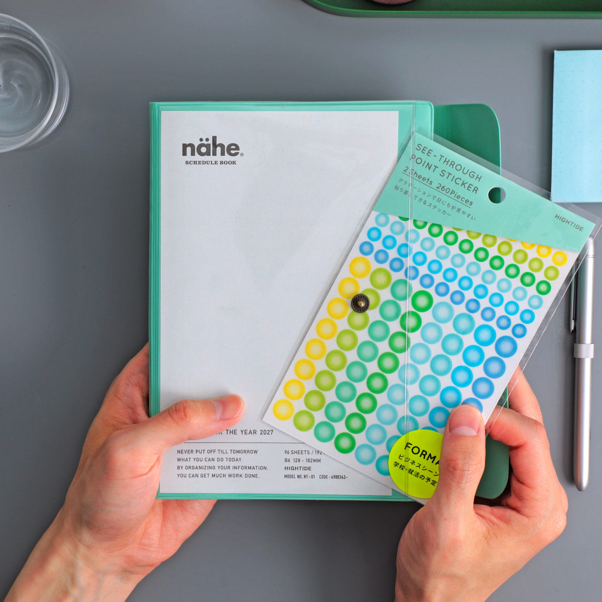 Hands holding a Mint Nähe B6 planner, its clear front pocket storing a packet of colorful dot stickers.