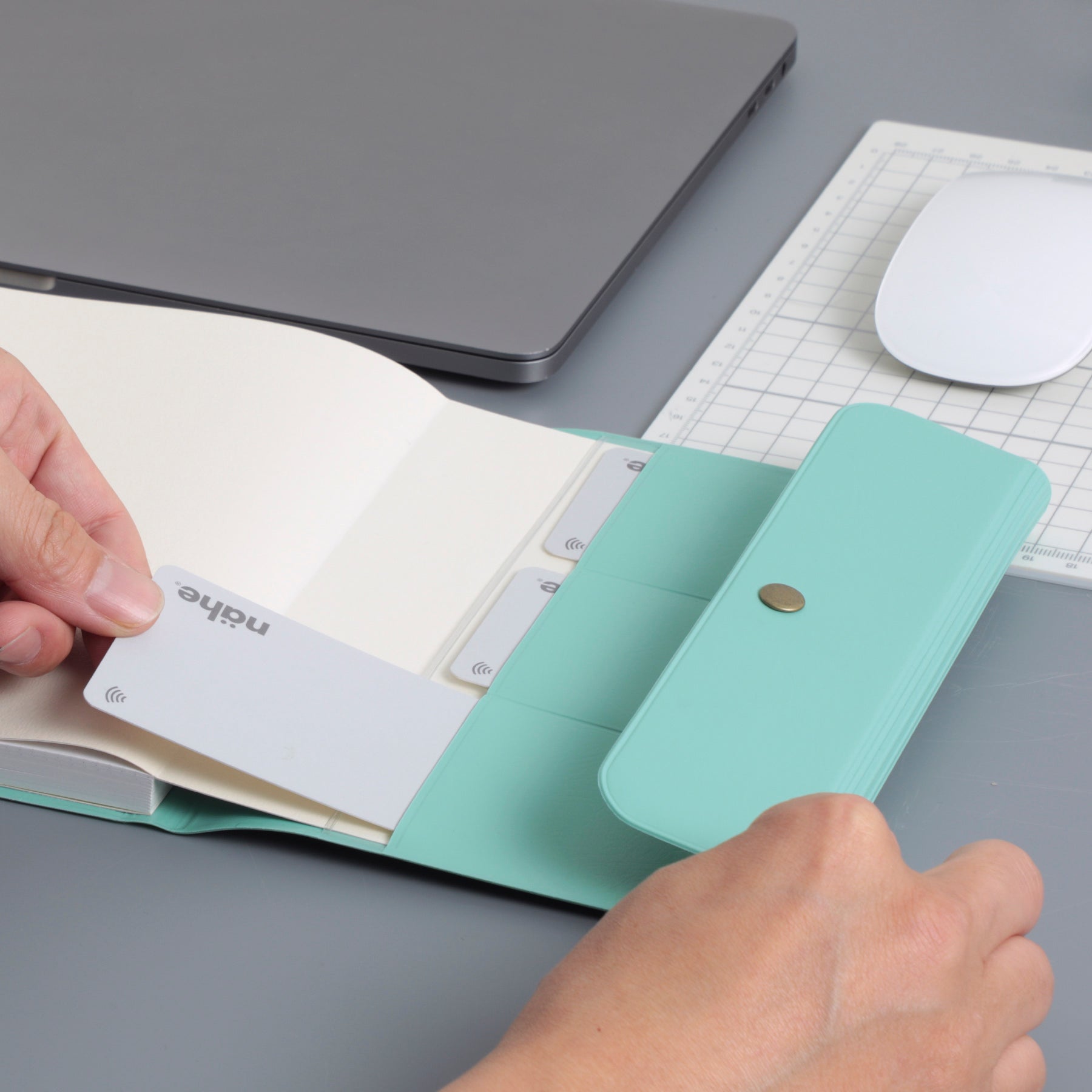 Hands slipping a card into the pockets of an open Mint Nähe B6 planner cover beside a laptop.