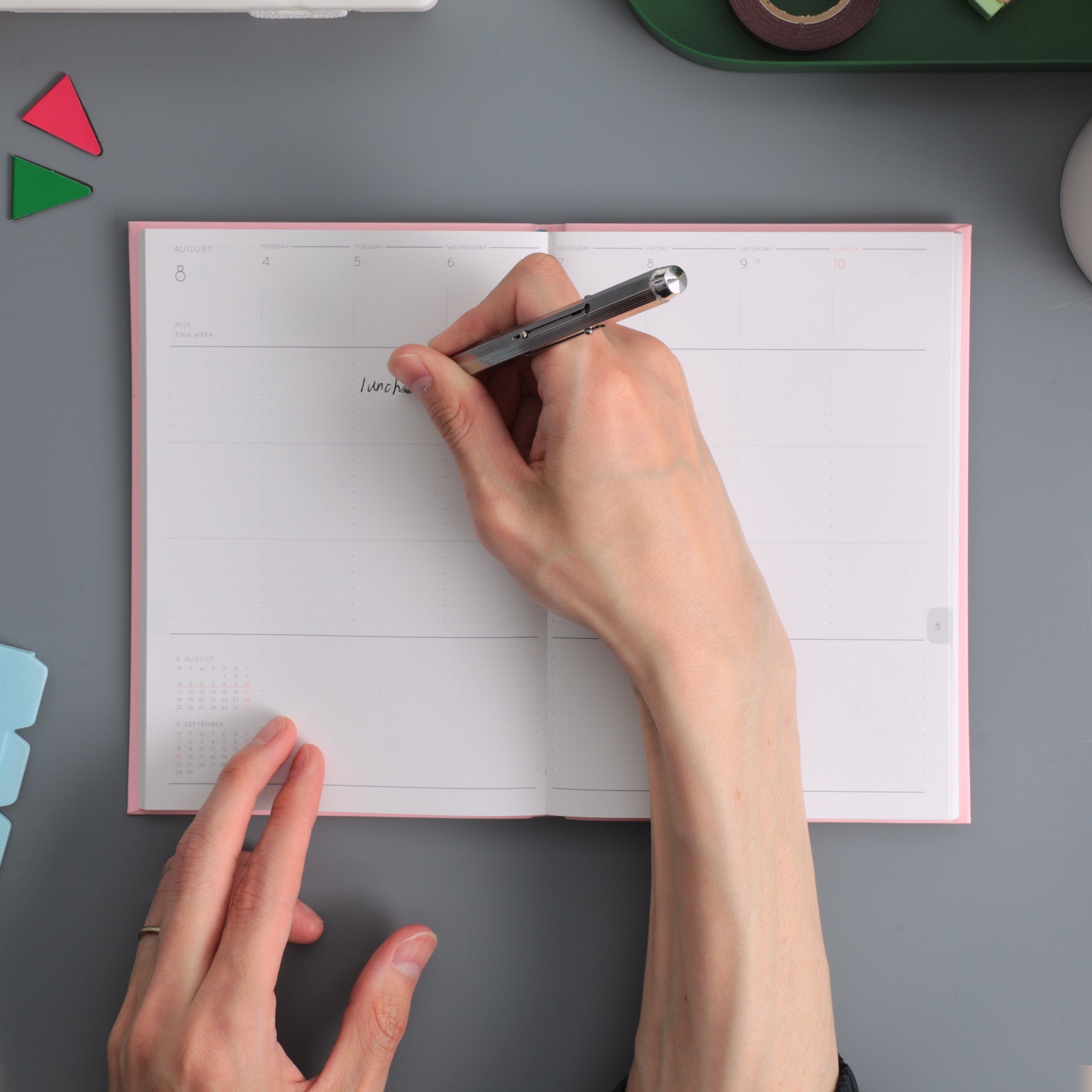 Hand writing with a pen in an open Pink weekly planner showing dated week spread on a grey surface.