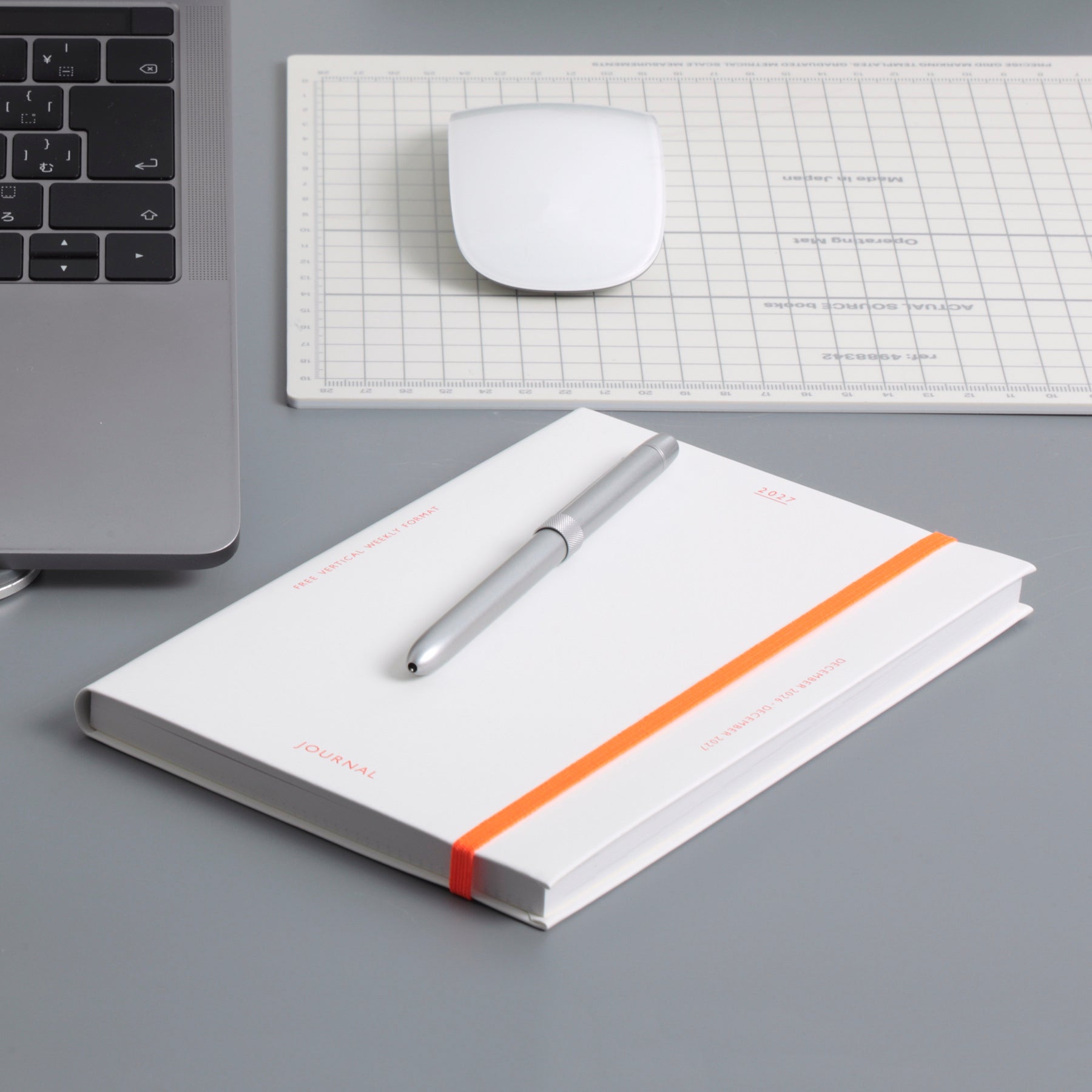 White hardcover planner with orange elastic band, silver pen on top, beside a laptop and grid mat.