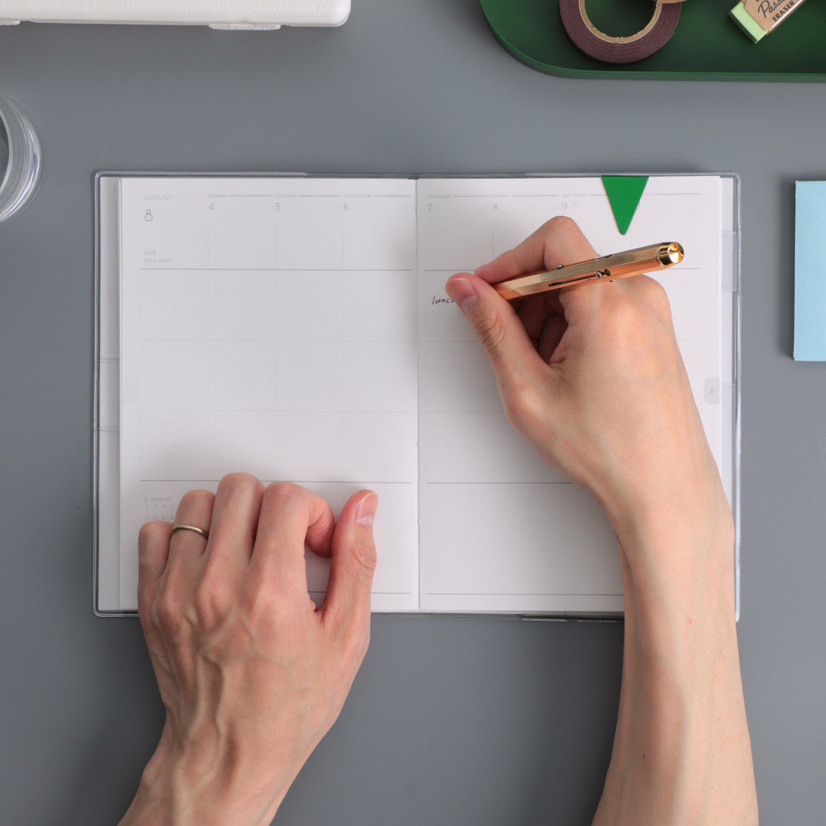Hands writing with a gold pen in an open weekly planner with blank ruled columns on a grey desk.