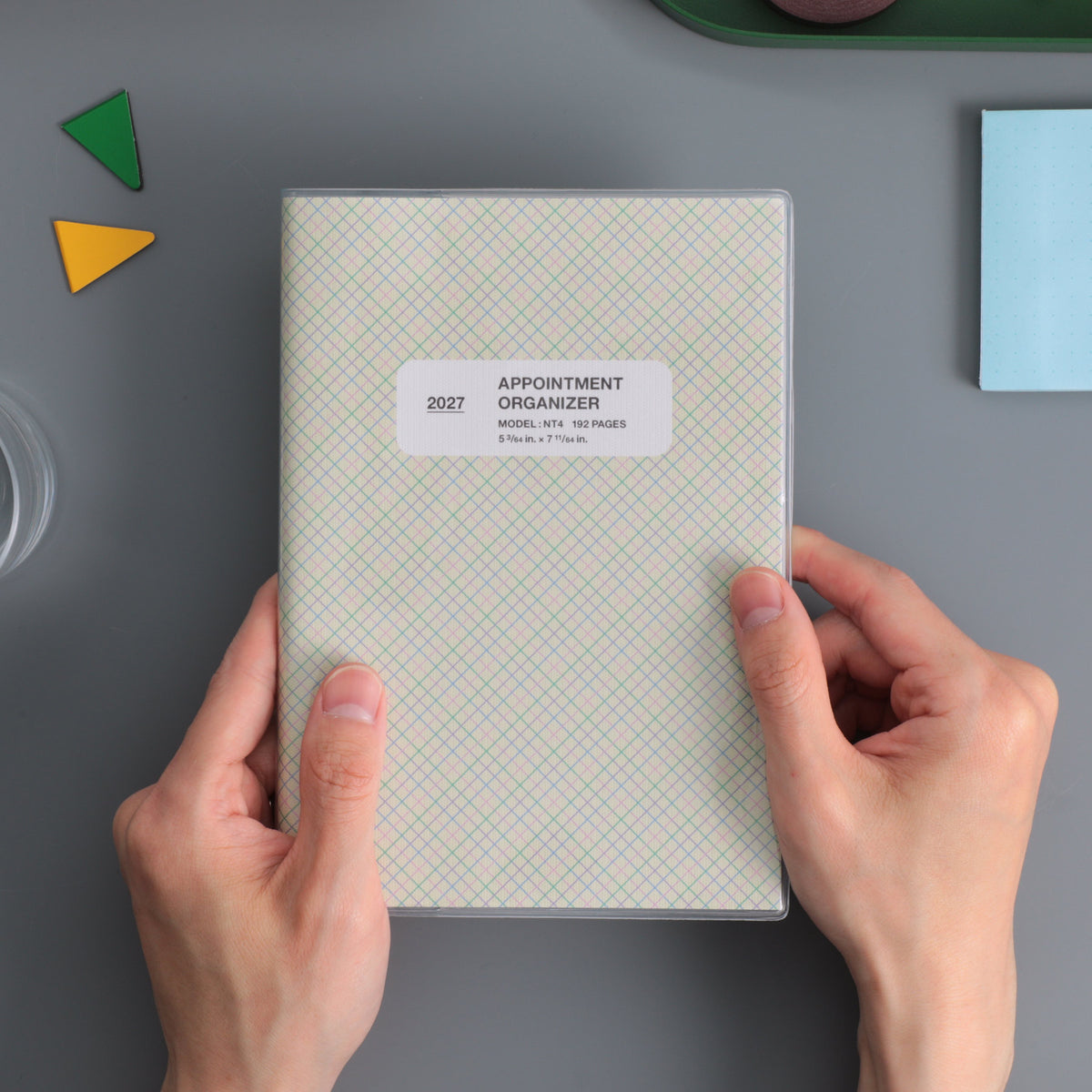 Hands holding a Hightide 2027 Reno B6 weekly planner in Pink check pattern with clear cover and label.