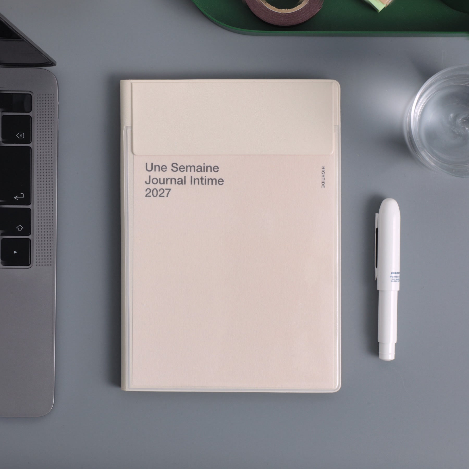 Ivory Hightide 2027 weekly planner with cover pocket, lying on a desk beside a laptop and white pen.