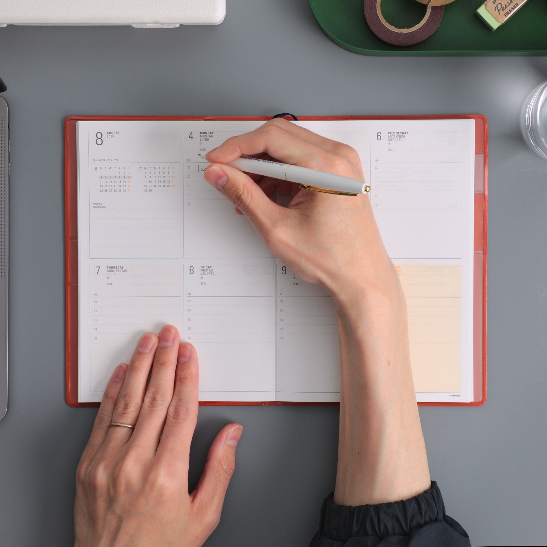 Hands writing with a pen in an open Orange weekly planner showing dated August pages on a grey desk.