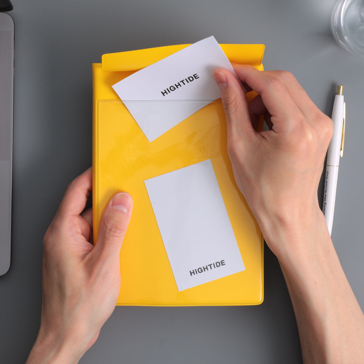 Hands slipping a card into the clear cover pocket of a Yellow Hightide planner, pen alongside.