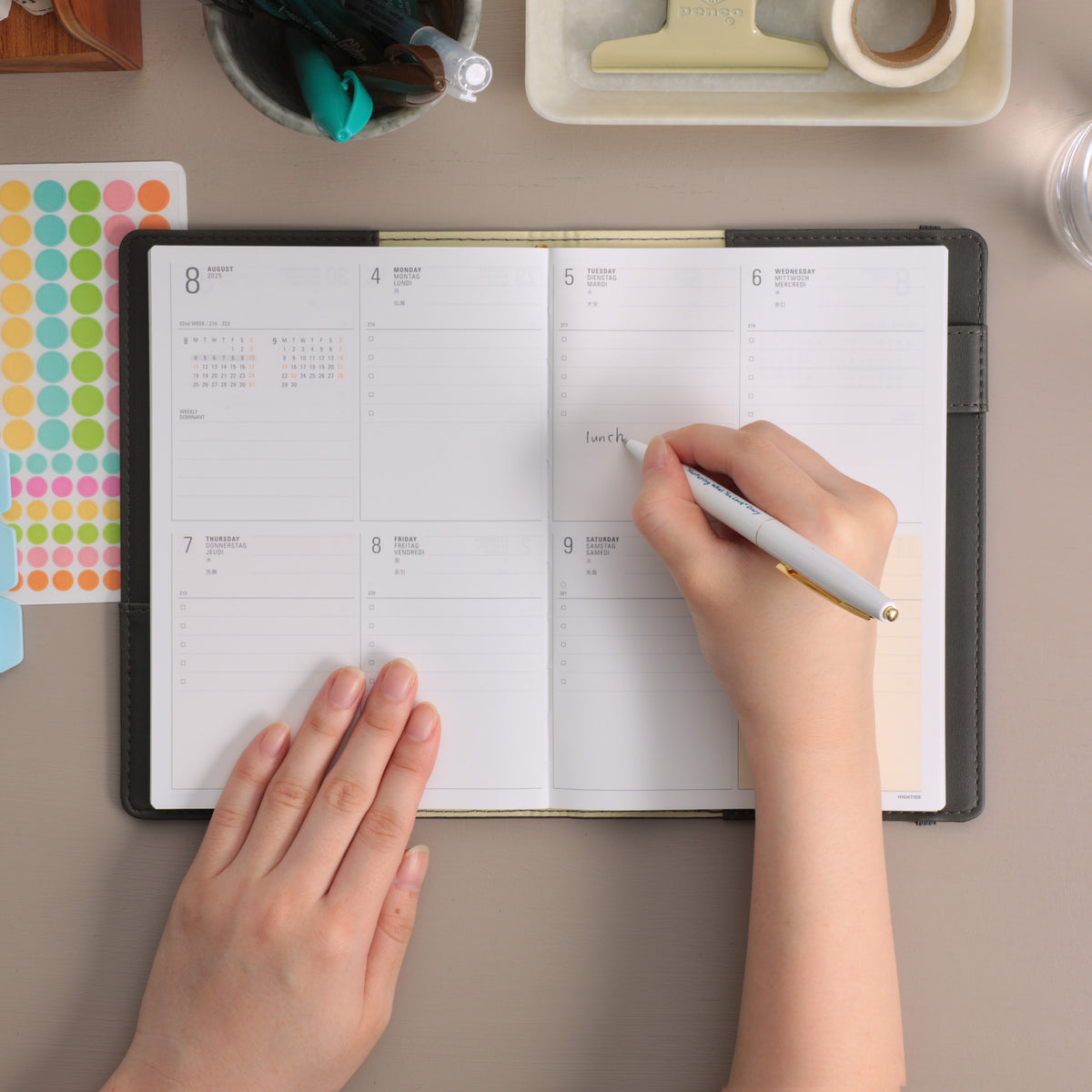 Hands writing "lunch" with a pen in an open Gray B6 weekly planner showing an August spread.