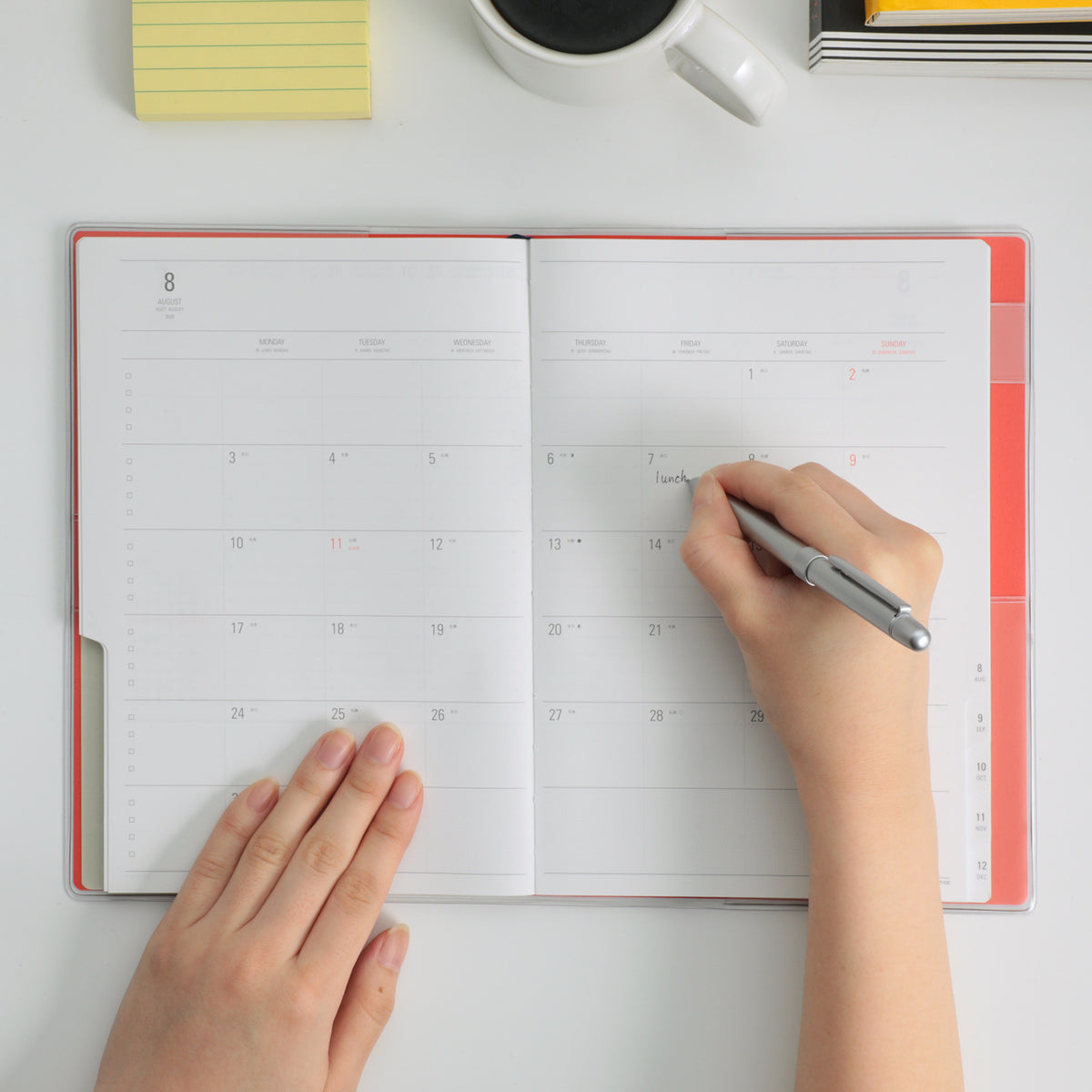 Hands writing with a silver pen in an open Nähe planner showing a monthly calendar spread, Red cover.