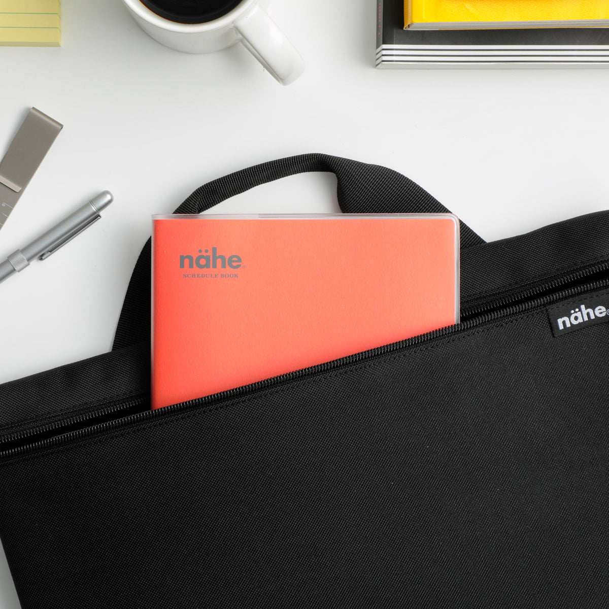 Nähe planner in Red slipped into the front pocket of a black bag, beside a coffee cup and pen.