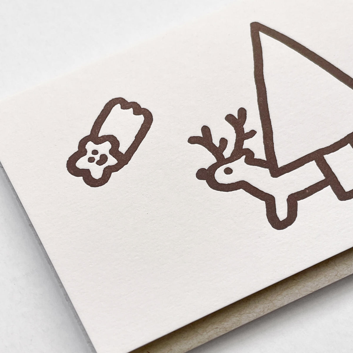 Close-up of letterpress card showing brown line-drawn reindeer, tree and star cookie on textured white paper.