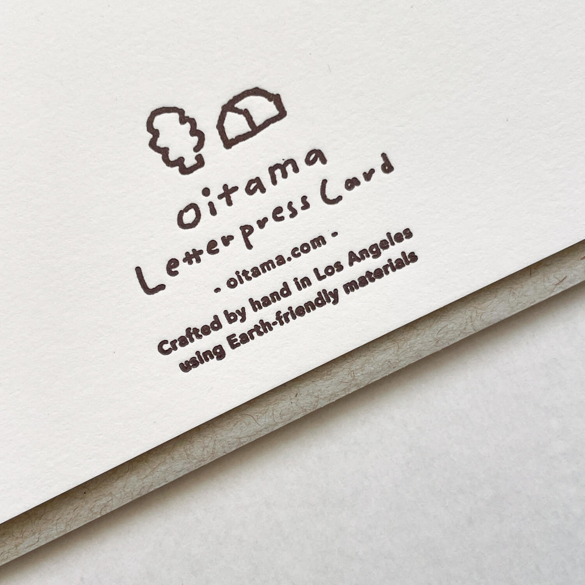 Close-up of the card's back showing the brown letterpress oitama logo and hand-printed text, with grey envelope beneath.