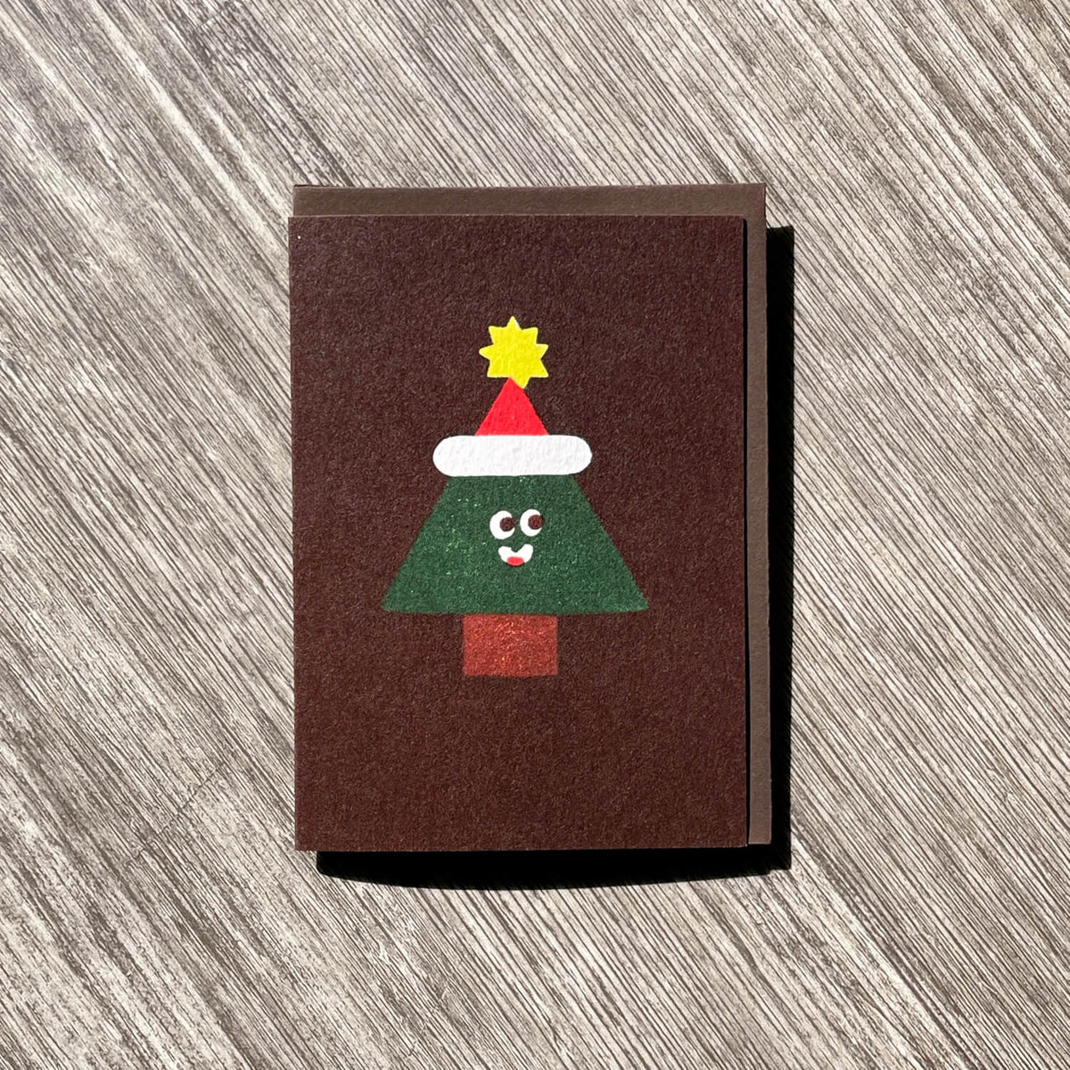 Brown greeting card with a smiling Christmas tree in a Santa hat and yellow star, on a matching envelope.