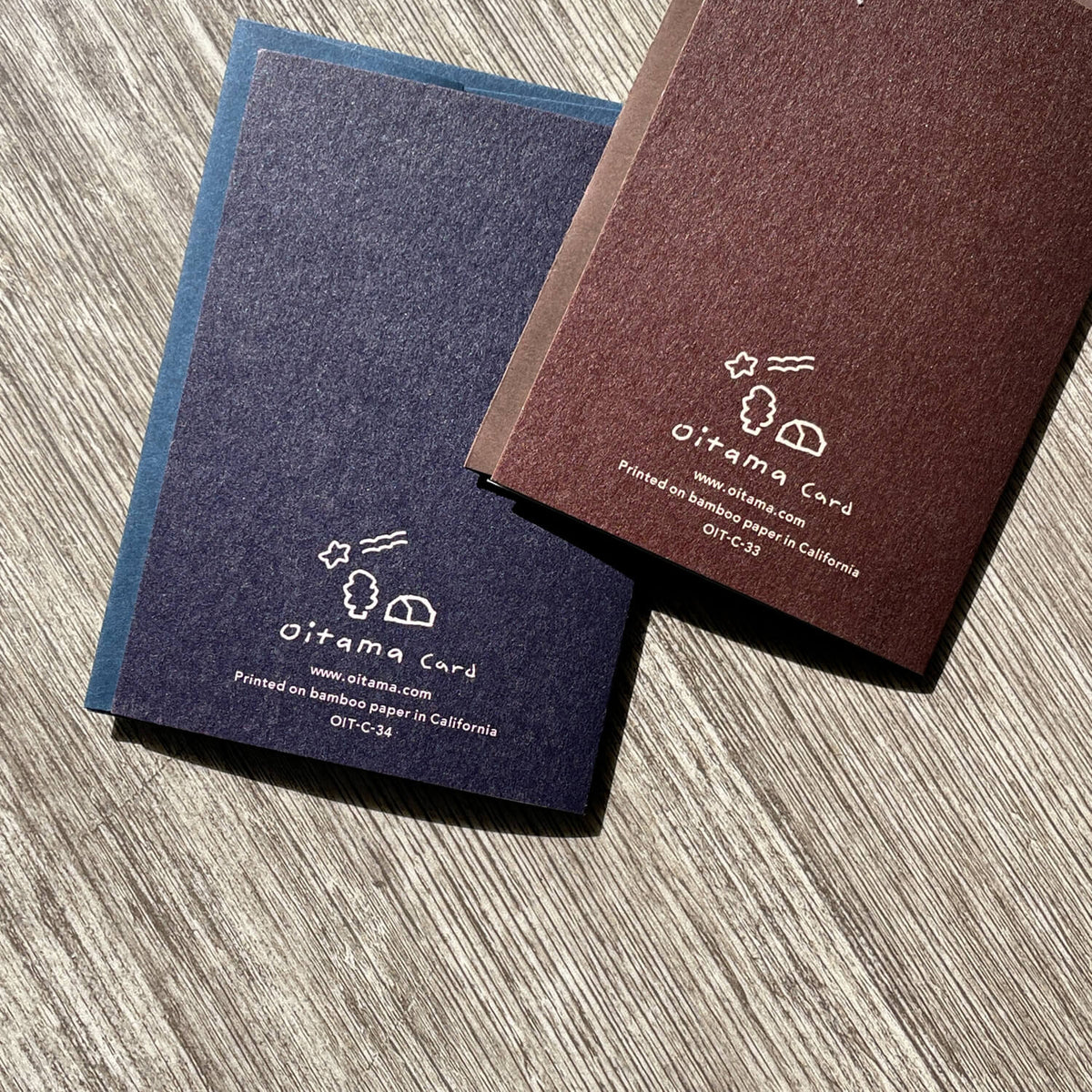 Backs of navy and brown Oitama cards on wood, showing white logo and bamboo paper printing details.