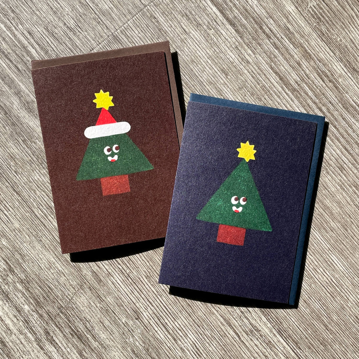 Two holiday cards on wood, one brown with a smiling Christmas tree in a Santa hat, one navy without hat.