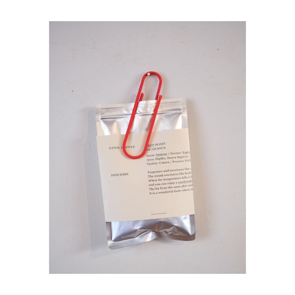Oversized Red clip holding a silver coffee bag with a printed label against a white wall.