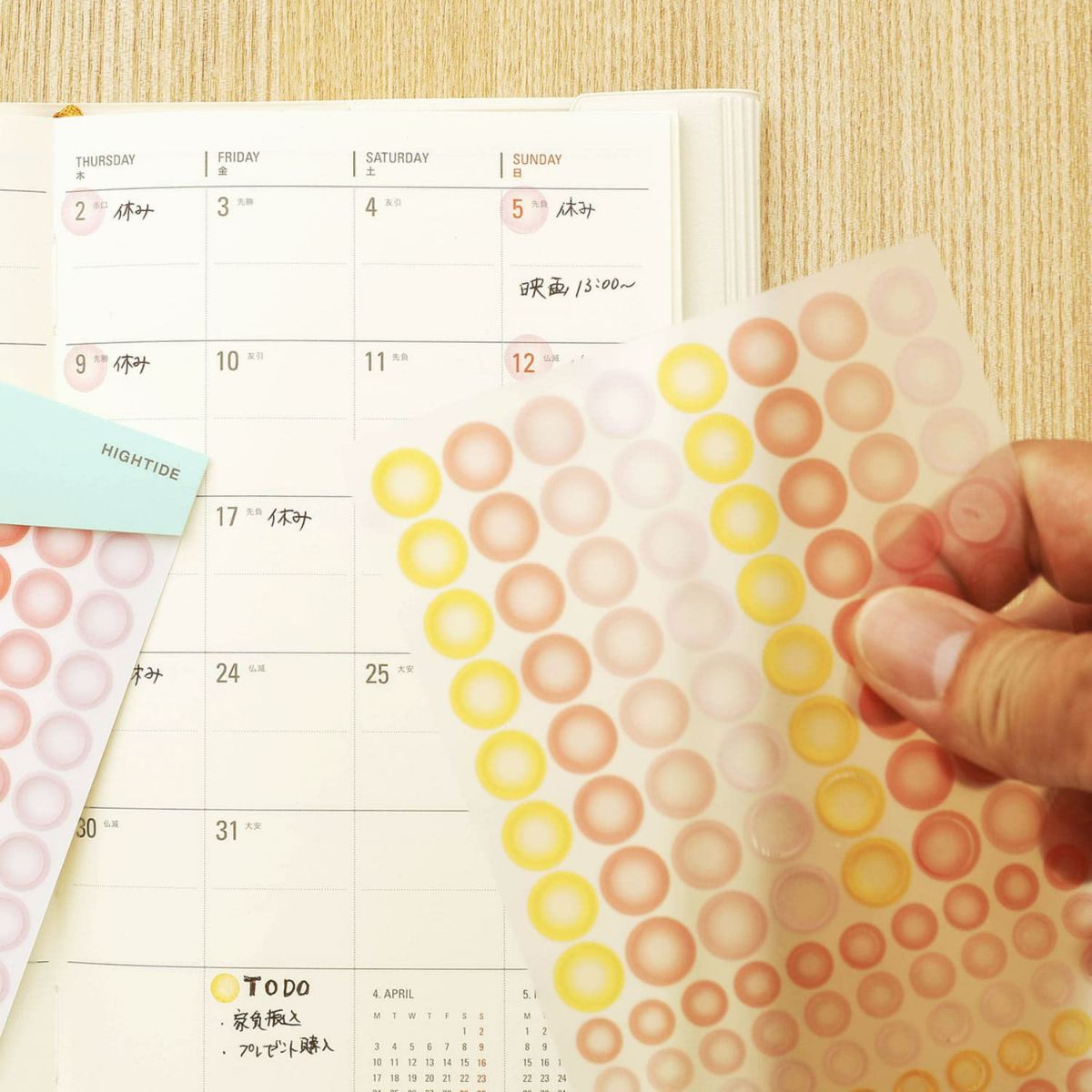Hand holding a sheet of see-through dot stickers in yellow, pink and coral above an open monthly planner.