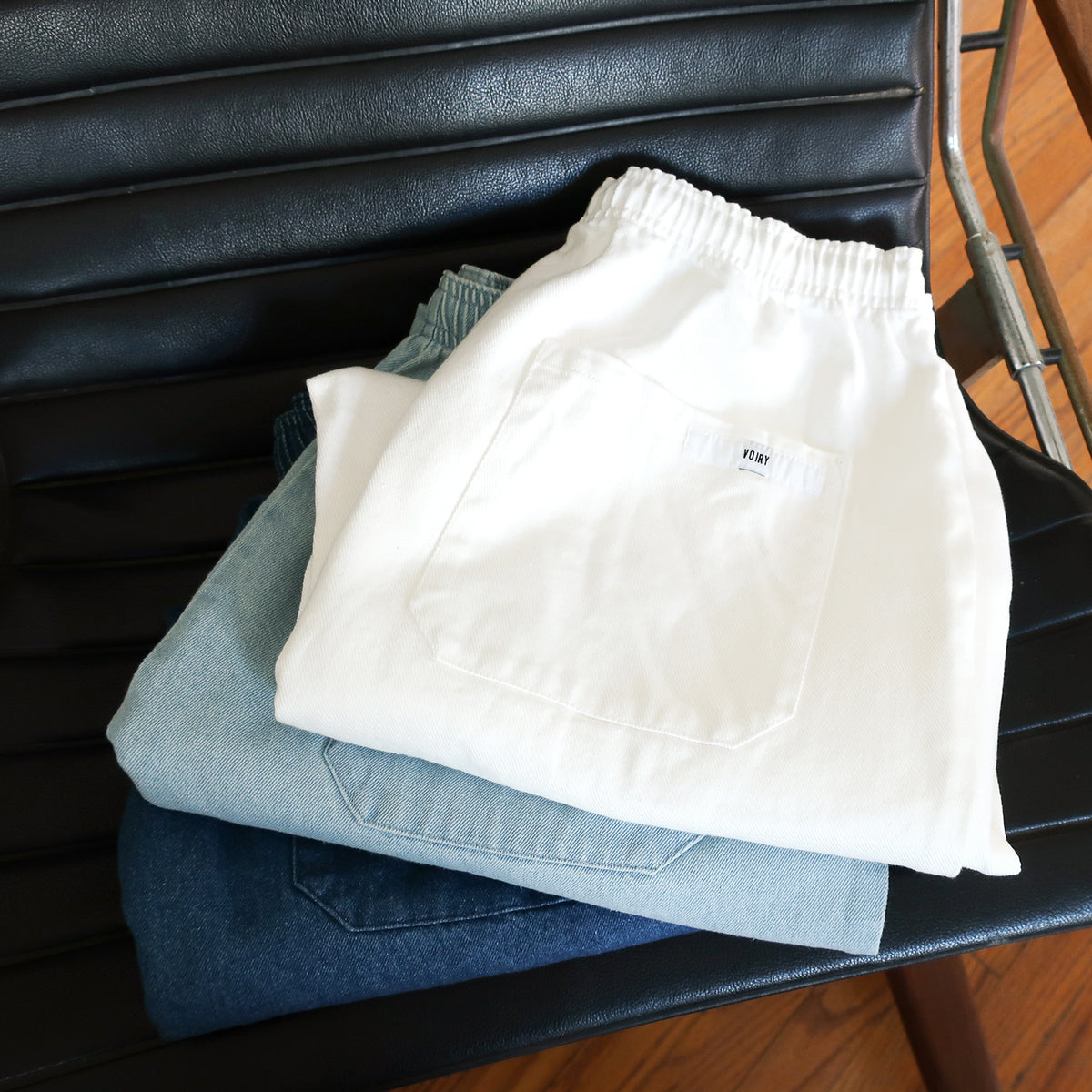 Voiry Sunday Pants folded and stacked on a black leather chair, White Denim on top with patch pocket and brand label