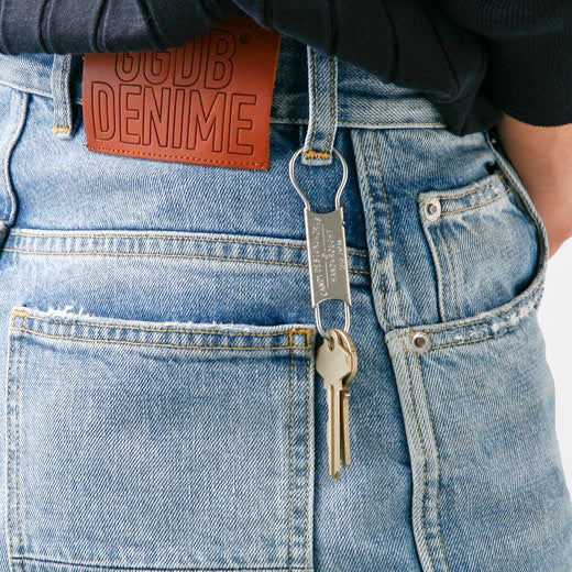 Stainless steel key ring with engraved tag and brass key, clipped to the belt loop of light blue jeans.