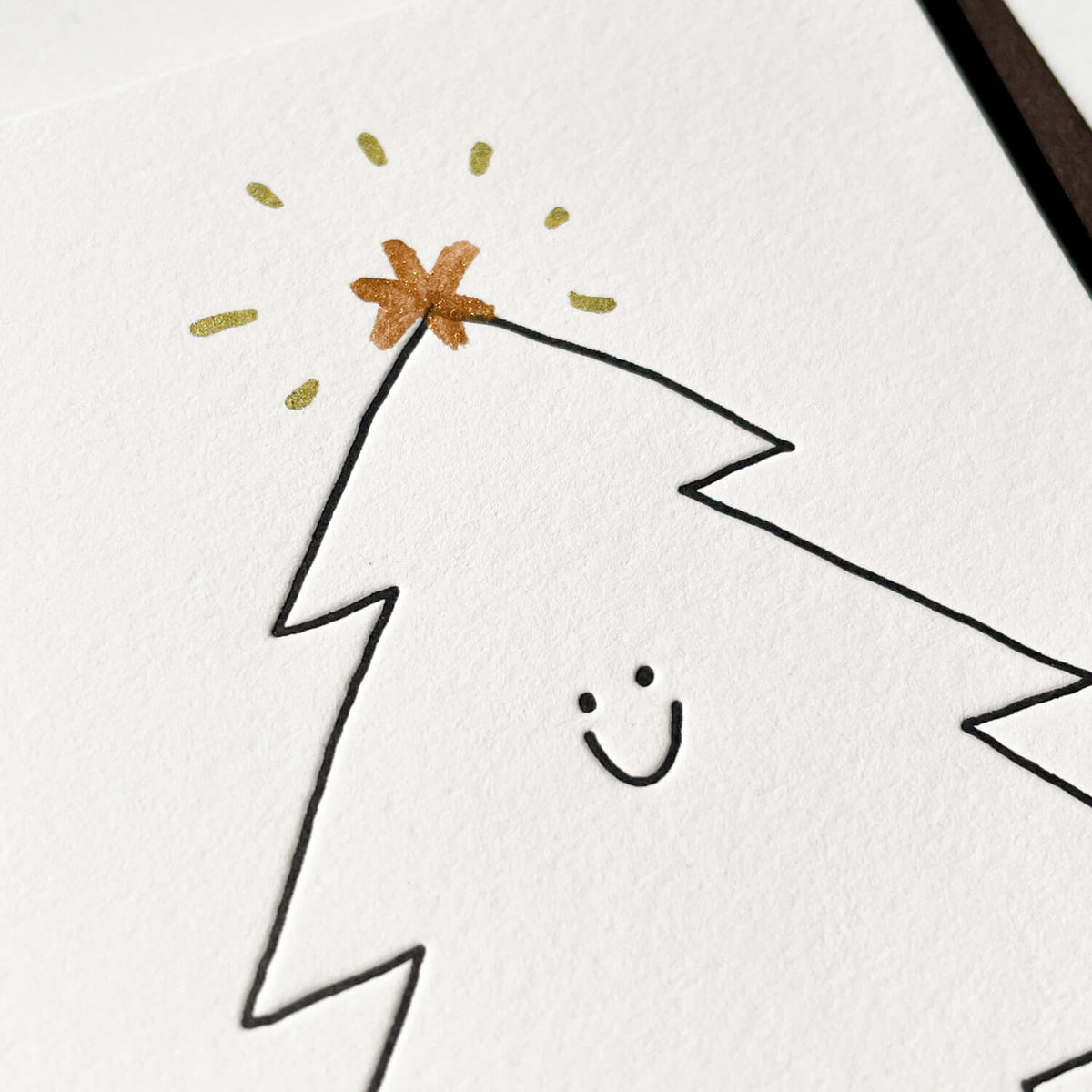 Close-up of a letterpress card showing a smiling outlined tree topped with a gold and copper star.