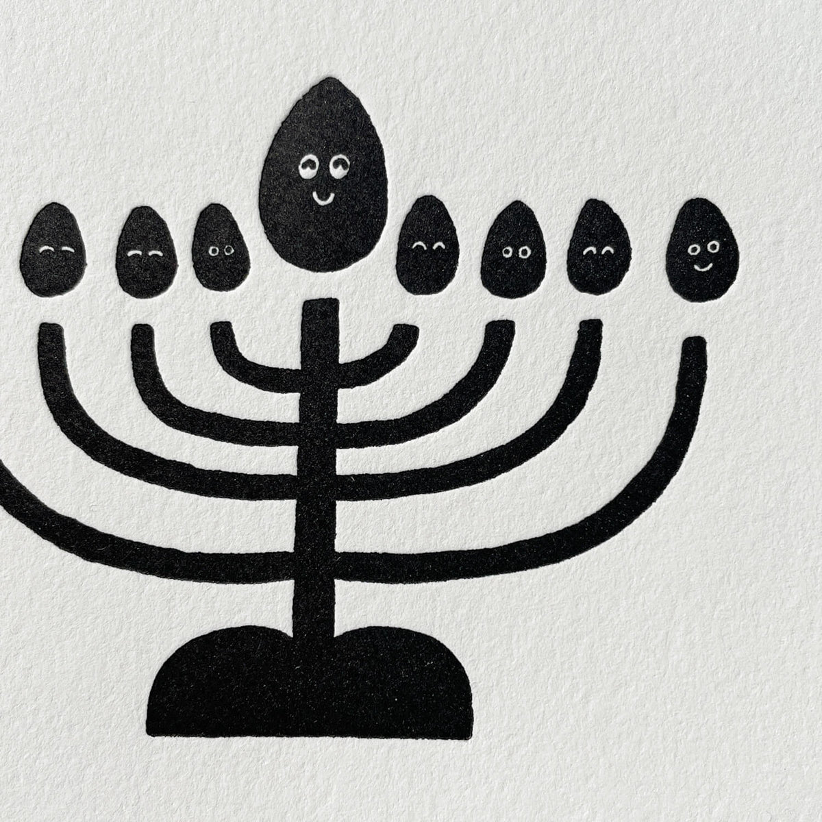 Close-up of black letterpress menorah illustration with smiling flame faces on textured white card.