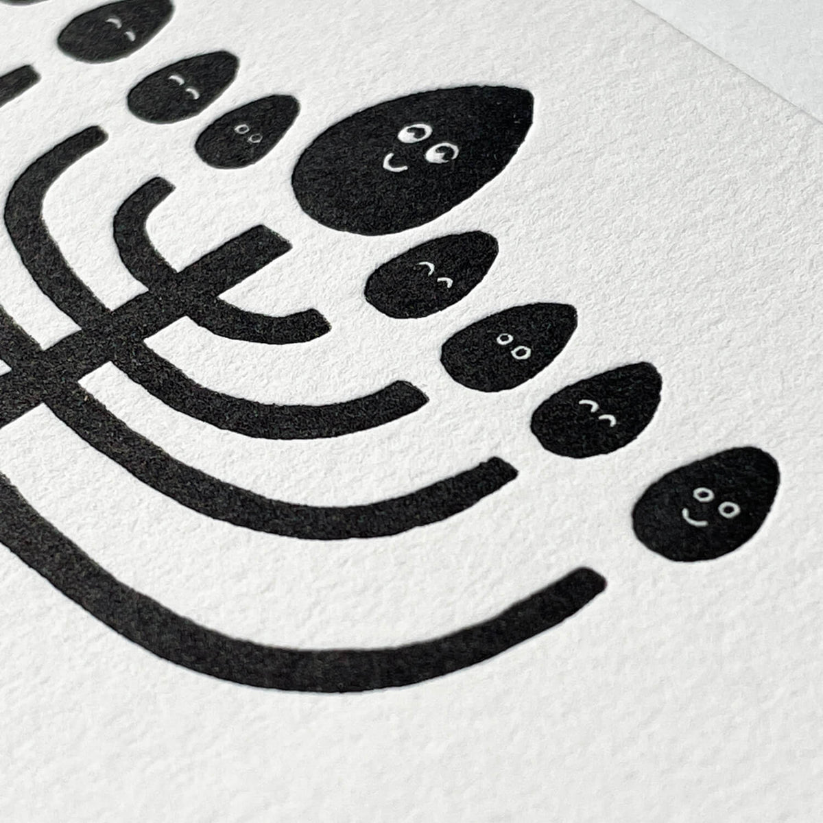 Close-up of letterpress card showing black menorah with smiling face flames pressed into textured white paper.