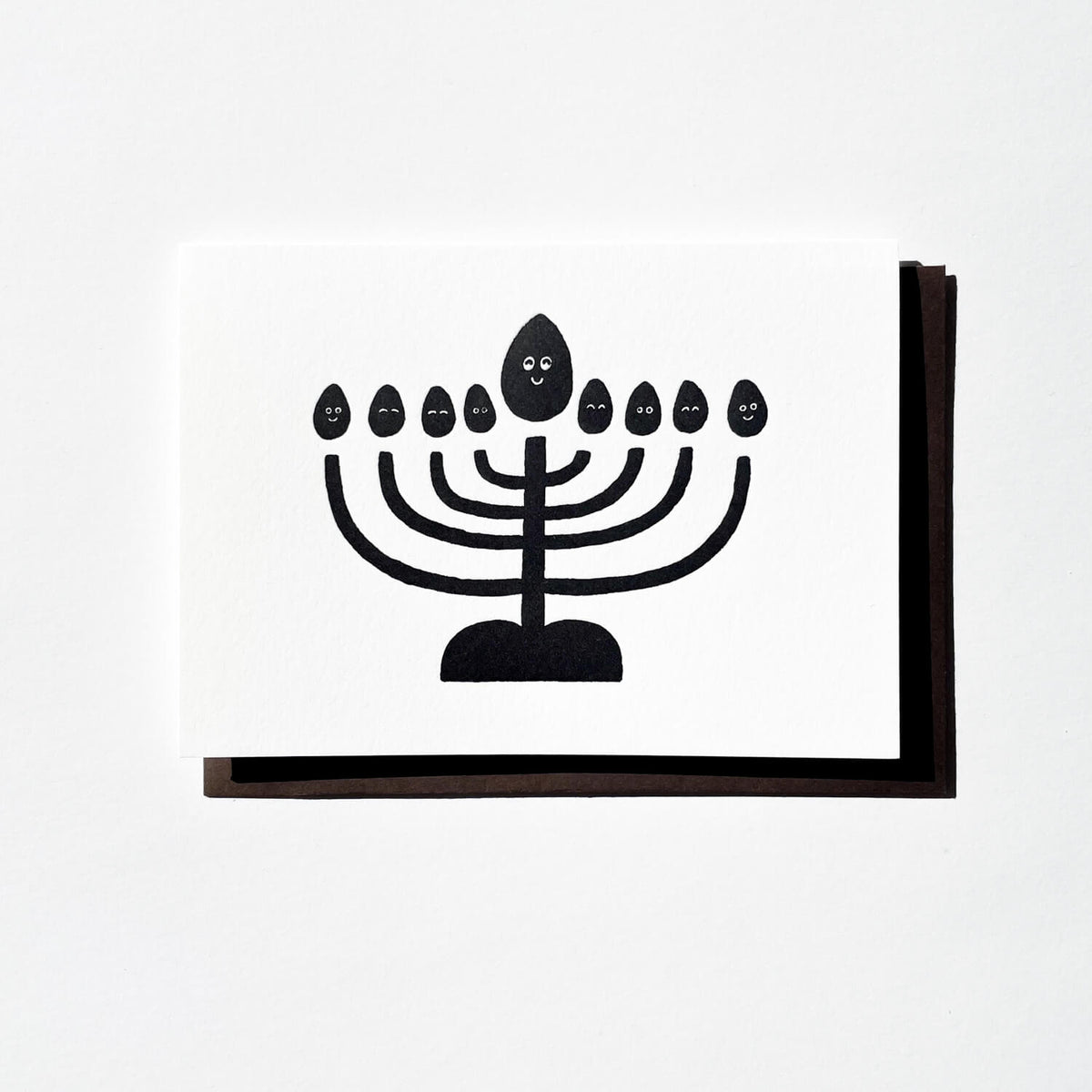 White letterpress card printed with a black menorah topped by smiling flames, with dark envelope behind.