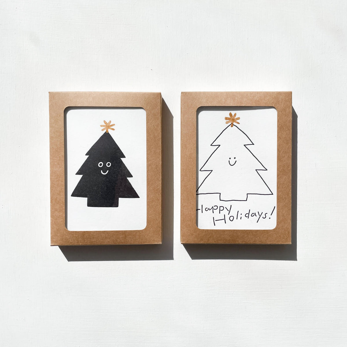 Two kraft boxes with window openings showing letterpress cards of smiling holiday trees and "Happy Holidays!"