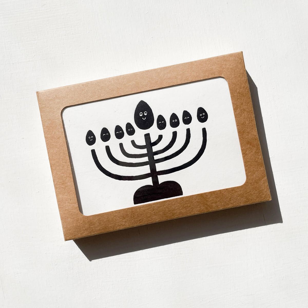 Kraft box with window showing a letterpress card printed with a black menorah with smiling flames.